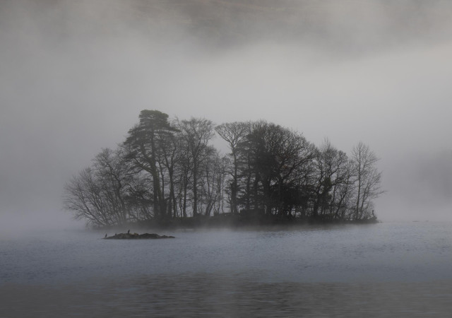 A tree covered island is shrouded by mist in the centre of a lake. In front of the island is a smaller rocky island with three black cormorant like birds. 