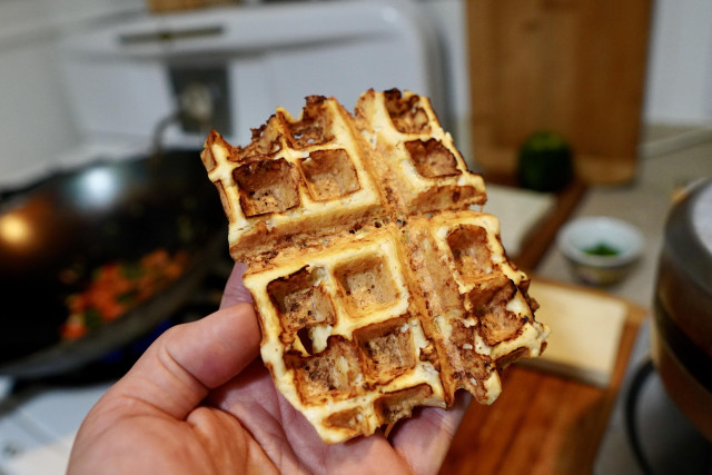 A hand holding what looks like a waffle, but is actually a piece of tofu. It has the same golden brown color and shape of a tofu. 