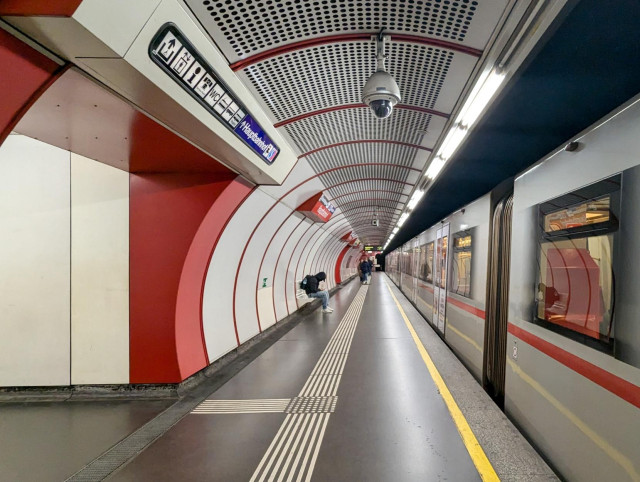 U-Bahnstation in Wien mit den typischen Designelementen wie weisse Panele mit und ohne Löcher, Ecken und Tafeln in der Linienfarbe und wunderbarer Helvetica-Typographie. Die Stationsrückeand ist halbrund gebogen, ein Zug Typ V steht in der Station.