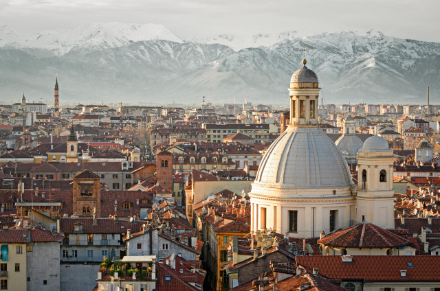 An aerial view of Turin, Italy, with a prominent domed building in the foreground and a dense arrangement of historic buildings with red-tiled roofs. Snow-capped mountains are visible in the background, highlighting the city’s scenic location at the foot of the Alps  