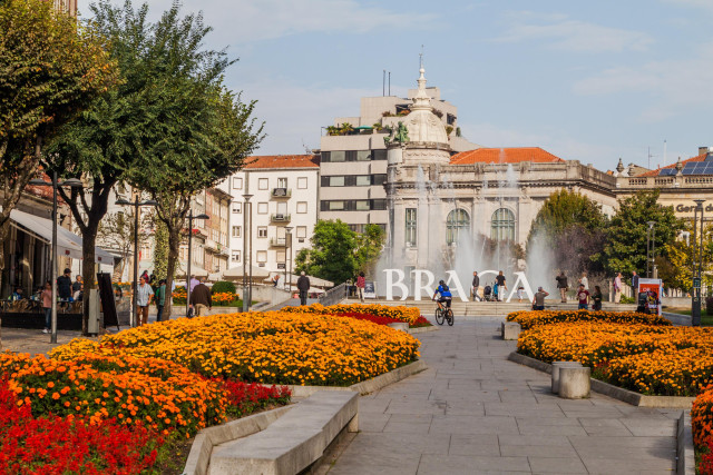 A city square with a large fountain in the centre, surrounded by people and cyclists. Orange flowers are in the foreground, with buildings in the background and a clear sky above.  