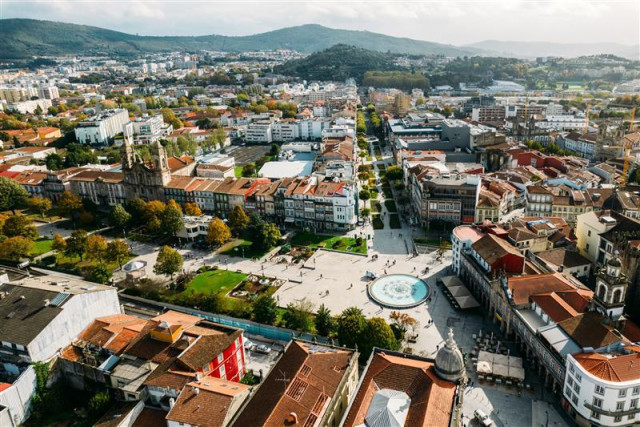 An aerial view of Braga, Portugal, showcasing a mix of historic and modern buildings, with a central square featuring a circular fountain. The city is surrounded by green hills, with tree-lined streets and parks visible, creating a vibrant, picturesque urban landscape 