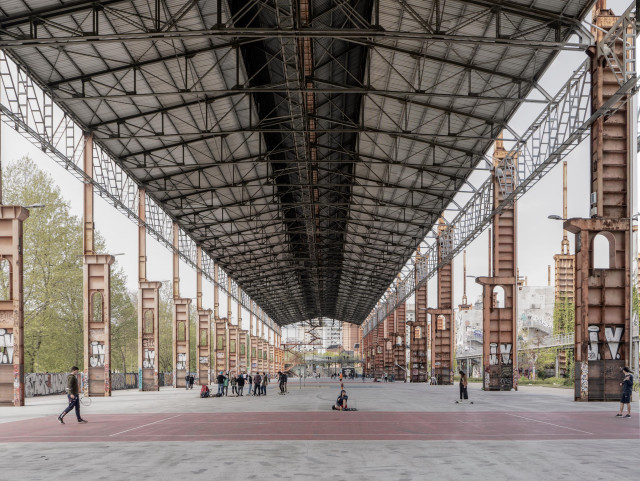 A photo of a large industrial-style roof with a repeating pattern of steel trusses and columns. Across the open ground below are people talking or practising sport. 