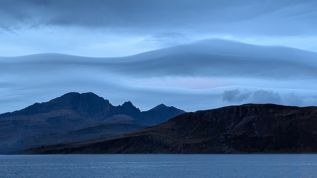 Wispy, wavey clouds sitting over a mountain, everything has a blue tinge as the light is cold just after sunset. 