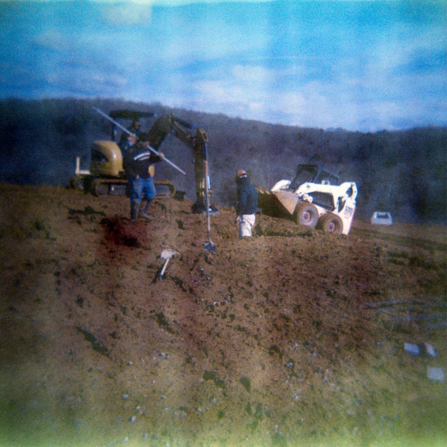 Workers and construction equipment onsite for a new building. The colors are close to normal after significant editing to restore the image, but there's bar of green discoloration along the bottom of the dirt pile they're standing on. 

There are also noticeable vertical "waves" extending across the photo.