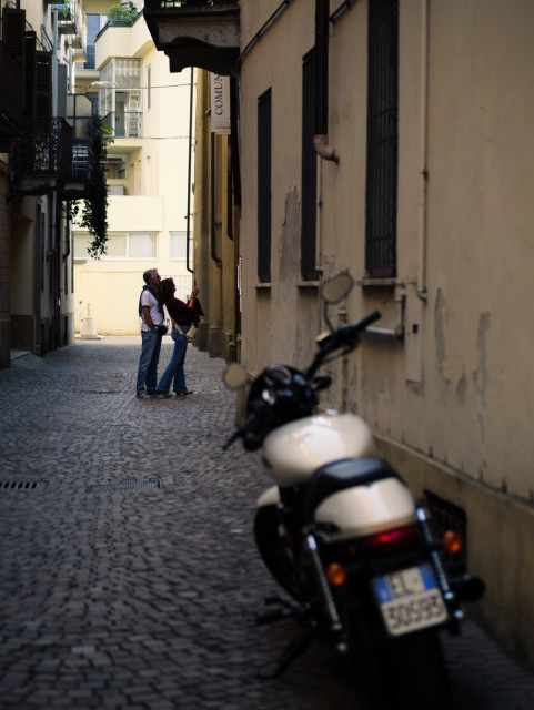 A couple look up at something above their heads in a narrow alley. In the near ground, an out-of-focus classic motorbike is parked.