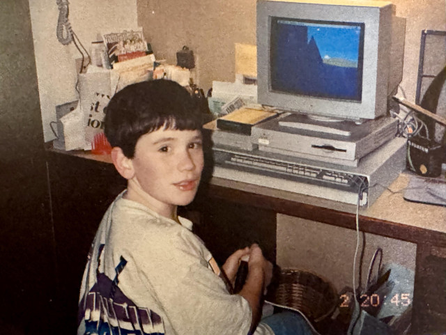 A young boy in a t-shirt sits at a desk with an old computer setup, including a monitor, mouse, keyboard CD drive, and modem. The background contains various items like papers and a phone.