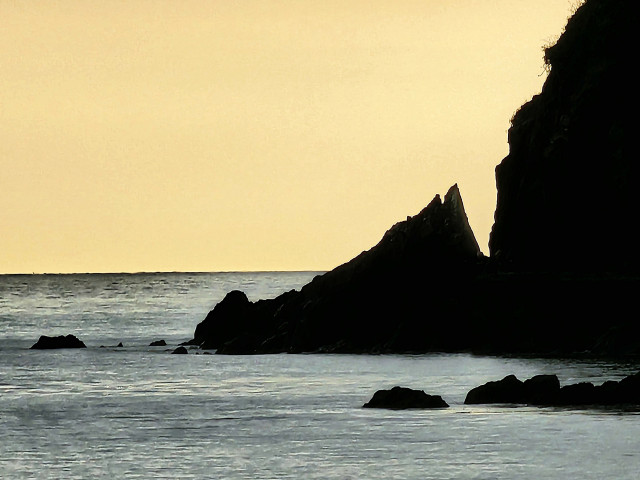 A silhouette of rocks against sea and sky. The rocks are to the right and the sky has a yellow cast.