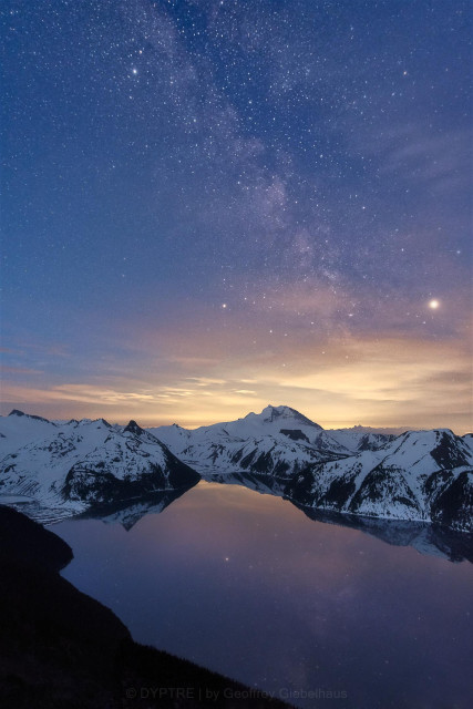 The core of the Milkyway sits above an imposing snow covered peak. It's reflected in the lake below. Yellow light pollution from distant city brightens the horizon.