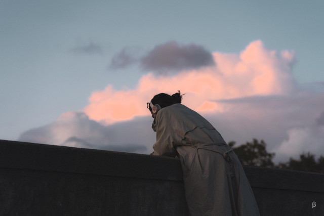 The image shows a person silhouetted against a dramatic sky with vibrant pink and orange clouds. The individual appears to be standing on a rooftop or ledge, gazing out at the sunset. The composition and lighting create a contemplative, introspective atmosphere.