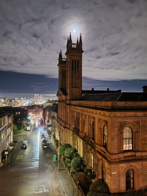 A photo of a tall sandstone building with a tower, at night, with a street on the left, and the sky above. The moon is bright and just above the tower, circled by many white clouds.
(Edited brick to sandstone)