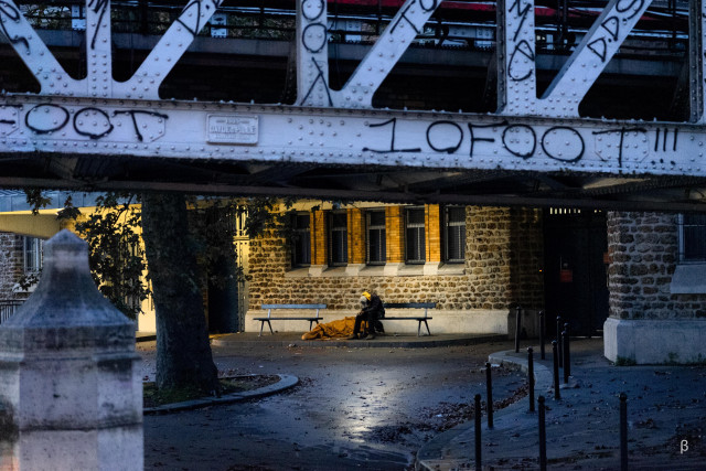 The image shows a nighttime urban scene under a large metal bridge structure. The bridge has intricate metalwork and the word "LOFOT" is visible on the side. Below the bridge, there is a stone building with illuminated windows, and a person sitting on a bench in the foreground. The scene has a moody, atmospheric quality with the mix of light and shadows.