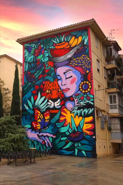 Streetartwall. On the outside wall of a four-storey building, a beautiful mural of a woman surrounded by flowers and animals has been painted in a naive style. The mural is painted in bright colors and shows the face of a young woman with closed eyes. She is wearing a turban as a headdress on which fruit is draped. She is surrounded by a jungle of green ferns and butterflies. Hummingbirds buzz through the picture and the head of an orange jaguar appears next to her in the jungle.
Info: The Theme was "Colombia". The artist wanted to represent freedom and coexistence by using some elements such as endemic species from Colombia like the jaguar, hummingbirds or butterflies. The female figure of Palanquera comes from a village called San Basilio de Palenque, the first slave-free village in Colombia.