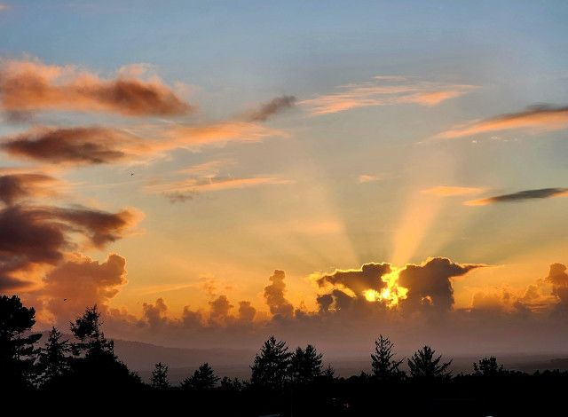 A stormy sky sunset with rays from the sun reaching up to the blue sky. The dark clouds around the sun are edged with gold. There is fog in the valley and dark tree tops at the bottom.