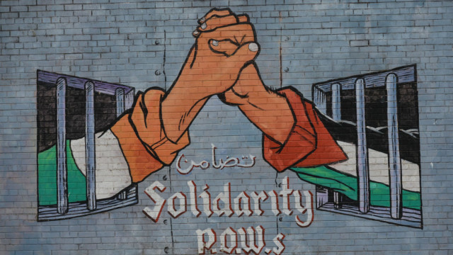 Muralmålning med irländsk och palestinsk hand bakom galler som möts, med texten "Solidarity P.O.W.s" (Solidaritet krigsfångar)