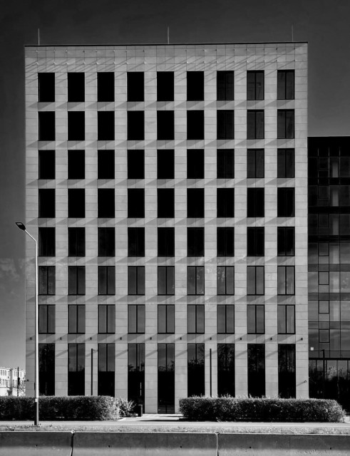 The black and white photo shows a modern building with a highly geometric, grid-like facade. The photo was taken from a tram line passing by. The building has a rectangular structure and is a mid-rise, about 8 to 10 stories tall. The surface is divided into neatly aligned, repeating squares with large vertical windows in each section. These windows are dark and reflective, contrasting with the lighter-colored material of the exterior walls, possibly concrete or stone. The shadows from the window frames give the building a three-dimensional, almost optical illusion effect, emphasizing the depth between each window.

In the foreground, there’s a small hedge along the base of the building. A streetlamp stands to the left of the frame, and a concrete barrier runs along the bottom edge. 