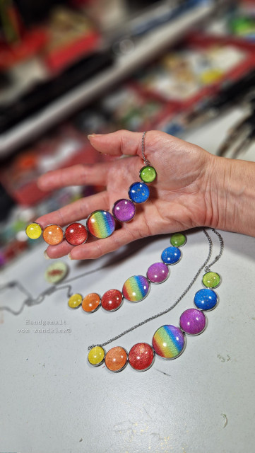 Halsschmuck mit sieben in Regenbogenfarben abgestuften Galaxie Motiven, aneinandergereiht alle an einer Halskette, an meinem Arbeitsplatz auf meiner Hand fotografiert