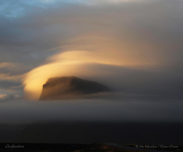 A photo of a beautiful mountaintop draped in misty cloud. Below, in the foreground, are scarves of mist above a dark ground surface. The peak of the mountain is peeking out between layers of vapour -above is a glorious crown of lenticular clouds tinted by the sunset into shades of orange, peach and grey. The sky above is clearing to cyan.