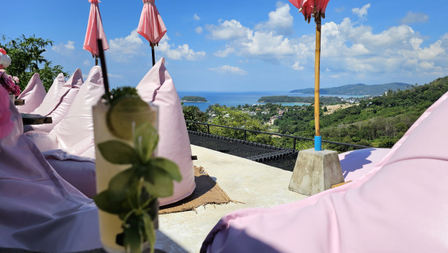 Das Bild zeigt eine atemberaubende Aussicht auf eine Bucht mit türkisfarbenem Wasser und grünen Hügeln. Im Vordergrund befinden sich mehrere rosa Sitzsäcke und Tische, die zu einer entspannten Atmosphäre einladen. Über den Sitzsäcken hängen rosa Sonnenschirme, die vor der Sonne schützen. Ein Cocktail mit einem Strohhalm steht auf einem Tisch im Vordergrund. Der Himmel ist blau und leicht bewölkt. Das Bild vermittelt ein Gefühl von Ruhe und Urlaubsstimmung.