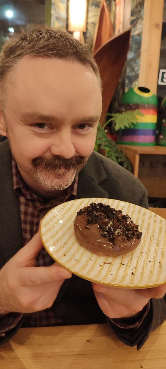 Picture of myself (mustachioed white man) holding a chocolate doughnut on a plate and grinning mischievously.