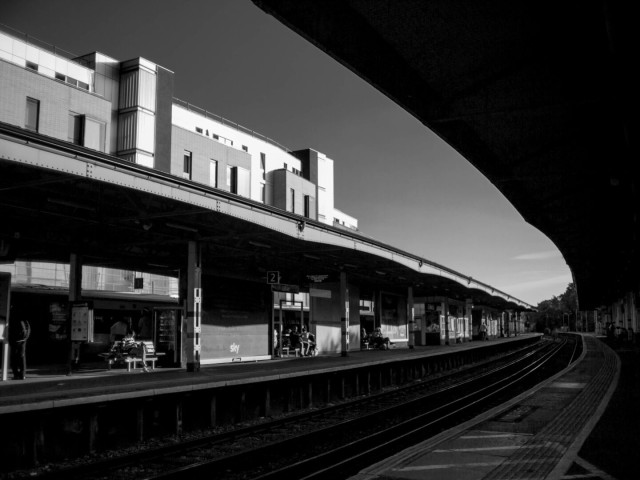 Monochrome wideangle photo taken from the end of a curved train station platform. The silhouetted canopy and platform edge on this side of the tracks take up the top and bottom right corners. Across two sets of tracks is an island platform with a canopy roof over several large advertising hoardings, between which sit people on benches. Behind the platform and the station itself is an apartment block made from several boxy rectangular sections. Above is the sky, clear except where it curves down to the horizon, where there's a couple of wispy clouds.