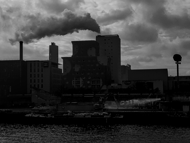 Black and white daylight urban photo.
on the dock of a river, chimney smoke, buildings in playful cloudy backlighting.