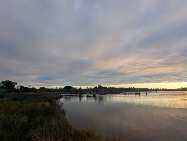 Photo of a calm morning sunrise over Harris Creek sun rising behind a low grey cloud deck with a clear horizon and tinting the lower clouds orange. Marsh is brown and ready to sleep for the winter. Creek water is calm and perfectly reflects the sky and clouds.