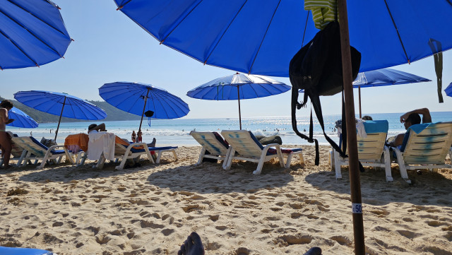 Blick auf den Strand und das Meer, am Strand stehen weiße Plastikliegestühle mit dunkelblauen Sonnenschirmen. 