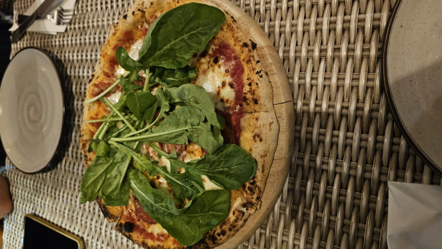 Auf dem Bild ist eine Pizza auf einem Holzbrett zu sehen. Die Pizza liegt auf einem Tisch mit einer geflochtenen Tischdecke. Die Pizza ist mit Tomatensauce, Käse und Rucola belegt. Der Rand der Pizza ist knusprig und leicht angebrannt. Neben der Pizza liegen ein Teller, Besteck und ein Handy.