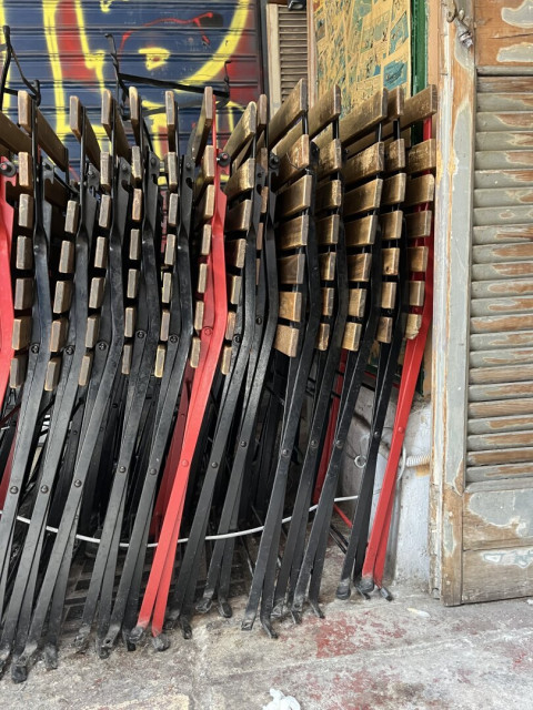 A line of folding tables with wooden slat tops folded against a wall under an apartment building. It’s a bar on the ground floor, and the outdoor space bounded by the support columns of the building above is a seating area. Most of the tables are black, three red, and it makes almost an abstract, a row of black metal lines with the odd re, spaced apart by the slats.
They’re chained to the concrete pillars and there’s an old wooden shutter decoratively by. There’s blue and red and yellow graffiti on the metal security shutter behind, but most of the image is folded tables