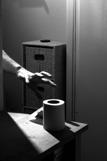 High contrast black and white photo of an arm stretching out of a room towards a toilet roll on a small table, just out of reach of the grasping hand. Behind the hand is a little wicker chest of drawers, on top of which is an infrared camera remote.