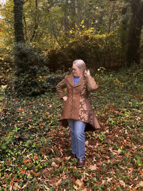 Me standing outside in autumn leaf covered grass, facing the camera and striking a silly pose, wearing a brown coat with a hemline that's shorter in the front than the back. The coat has vertical panels that are made of different fabrics, some print and some solid, all in different shades of brown.