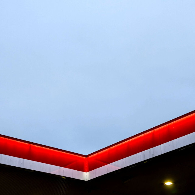 Photo looking up at a corner of the flat roof over a petrol station. There's a certain ambiguity due to lack of context, but it's an interior corner, most of the image is blank grey sky. At the bottom the edge of the roof is lit from under a small lip running around the top. The edge is otherwise a broad red band over a small white one. In the darkness underneath, there's a single small square light close to the bottom right corner. It's pretty dirty.