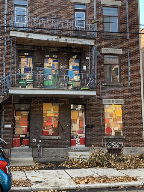 An urban building with multiple brick walls features several doors that are boarded up with colorful pieces of wood and cardboard. The upper balcony has decorative railing, and fallen leaves are scattered on the ground.