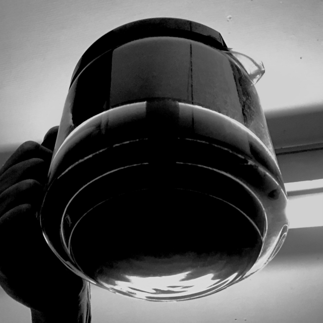 Black and white photo. Looking upward at the bottom of a half filled glass container with coffee. It is being held in place by a hand seen on the left side of the frame.