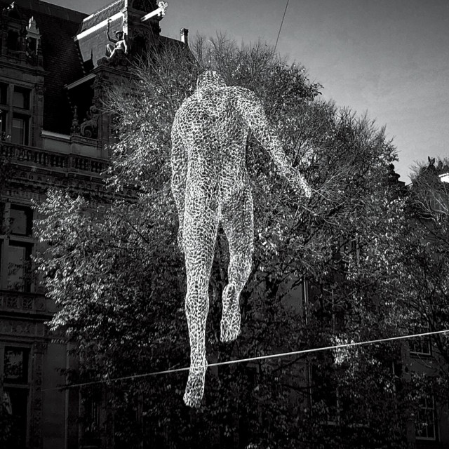 Black and white photo of a see-through wire model of the male form hanging above an Amsterdam canal with traditional old Dutch buildings and tree with autumn colours in the background against a blue sky.