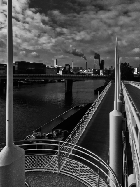 Black and White daylight phone photo.
From a bridge a view of the city quietly swallowing the river in a typical autumn sunny afternoon manner.