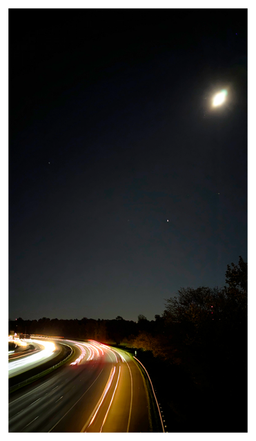 Saturn-shaped moon at top right over four lanes of freeway traffic, vehicles leaving light trails.