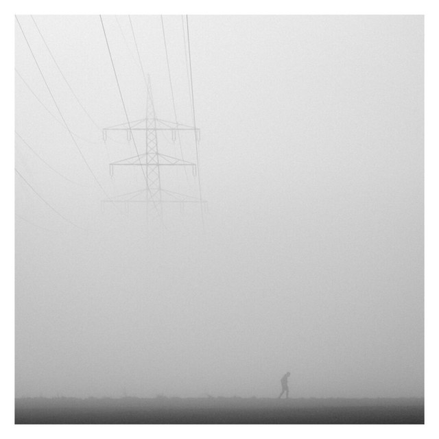 This square black-and-white photograph with a white border captures a foggy scene characterized by minimalism and contrast. A tall power line tower is positioned on the left side of the frame, its structure partially obscured by the dense fog, while its delicate wires stretch diagonally across the upper left corner, gradually fading into the mist. In the lower right corner, the silhouette of a solitary person walking adds a sense of scale and evokes a feeling of solitude. The flat, dark ground contrasts sharply with the light, misty atmosphere above. The white border emphasizes the clean, minimalist composition, enhancing the quiet and contemplative mood of the image.