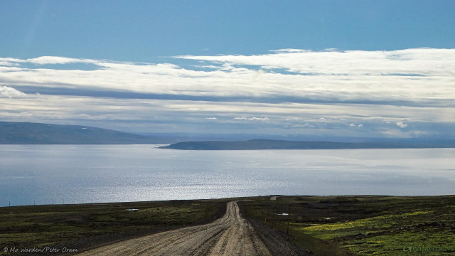 A photo of a landscape containing a large body of water. The foreground is a grey dirt road through dark grassland, puddles of standing rainwater are dotted throughout. Beyond that is the shining water of a huge fjord beneath a lightly clouded cyan sky. Heavier cloud is visible on the horizon, just above distant misty mountains.