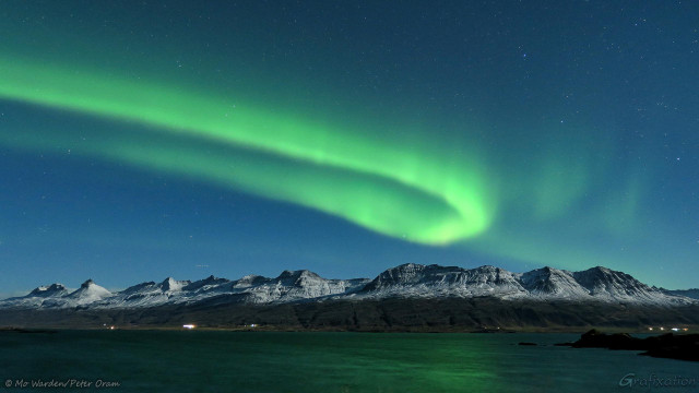 A photo of the night sky full of stars, with a beautiful green hook of aurora above moonlit mountains. The foreground is a fjord reflecting the strong green of the Northern Lights. On its shore are the lights of buildings. On the right, in the sky, wisps of faint aurora are visible and, on the left, a trail of dots left by the blinking light of an aircraft.