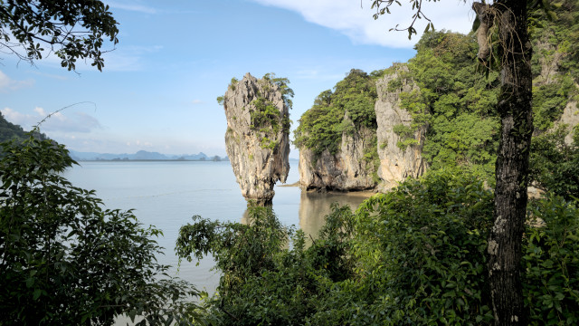 Das Bild zeigt eine beeindruckende Karstlandschaft mit einer schlanken, fingerförmigen Felsformation, die aus dem türkisfarbenen Wasser ragt. Im Hintergrund sind weitere, bewaldete Kalksteinfelsen zu sehen. Der Himmel ist blau und wolkenlos, was die tropische Atmosphäre unterstreicht. Das Bild vermittelt Ruhe und Schönheit und lädt zum Träumen ein.