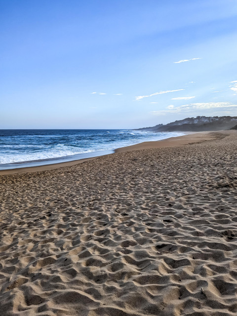 A sandy Pennington Beach, KwaZulu-Natal with a clear blue sky, hazy horizon and dark blue sea.