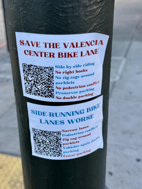 Two stickers on a post. One says "Save The Valencia Center Bike Lane" the other says "Side Running Lanes Are Worse". 

Both have a QR code and different bullet points supporting their argument. They are in a very simple font and design with a mix of red, blue, and black text. Not very eye-catching. 

Here are some of the bullet points: "SAVE THE VALENCIA CENTER BIKE LANE, Side-by-side riding, No right hooks, No zigzags around parklets, No pedestrian conflict, Preserves parking, No double parking."