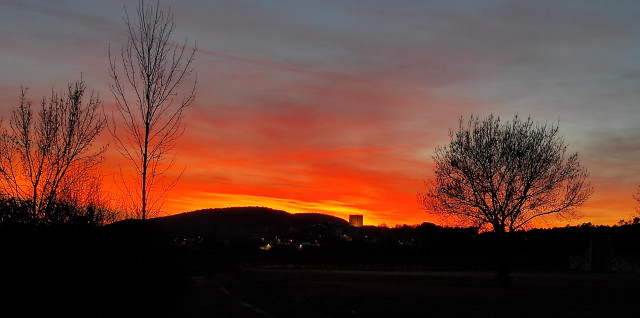 The sun is setting behind a hill and the sky is ablaze. There are the shadows of trees in the foreground 