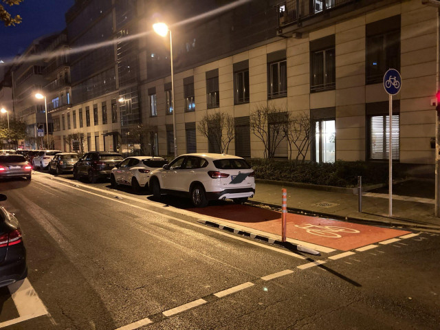 Straße in Frankfurt a.M. mit rot markierten und baulich abgetrennten Radstreifen, der von einer langen Reihe von parkenden Autos belegt ist. 