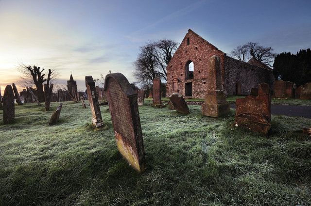 Photo of frosty grass around old gravestones with ruins of priory behind.