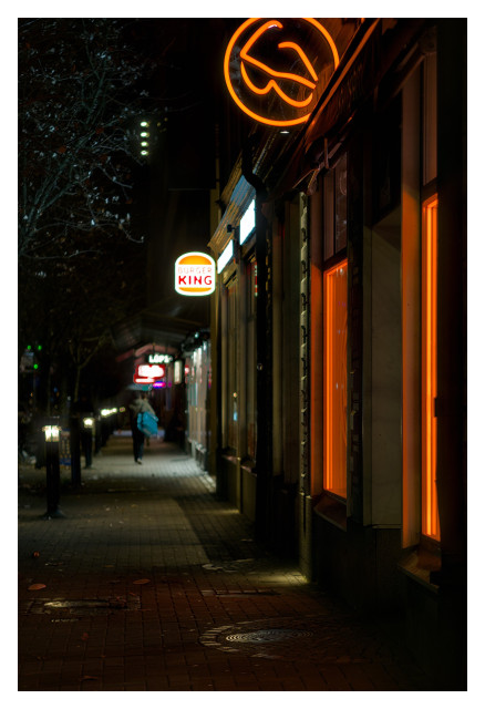 En trottoar i kvällsmörkret lyses upp av neonskyltar på höger sida. Först en orange Synsam, sedan Burger King och de senare är för suddiga att utläsa. En man med en stor blå väska över axeln går bort över.