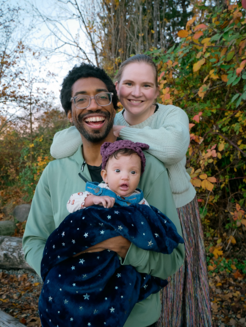 An outdoor family portrait of a seated Black man holding a baby, with a nonbinary white person leaning on his shoulders.