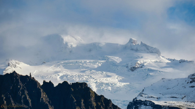 A photo of a mountaintop under a cloudy sky with large cyan patches. There's strong sunlight from the right. The foreground is uneven and dark rock ridges, bare of snow and ice but with unusual vertical fingers of rock sticking up from the edge. Behind this is a wide expanse of cracked and sculpted ice, clean and white with blue shadows. In the centre of the shot is the pinnacle. On the right it is an unusual twin stump of rock, almost like a small butte with a distinct ridge running away from it. On the left is a humped mound, and this appears to be the highest point.